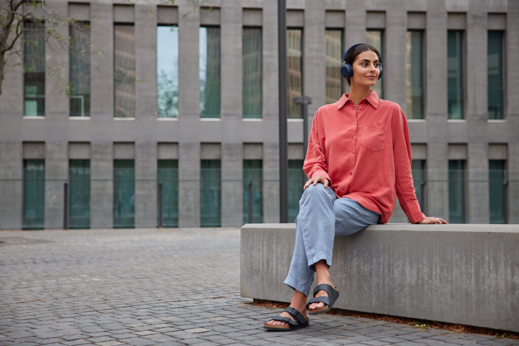 pleased thoughtful woman with dark hair wears comfortable casual clothes looks away listens music in headphones poses on stone plate near modern building. people lifestyle and hobby concept.